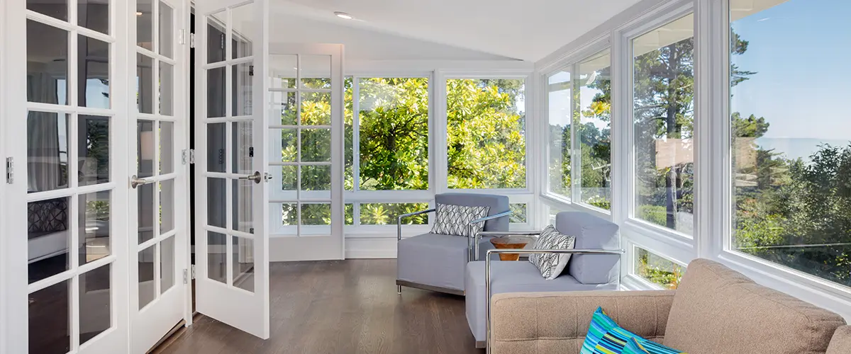 Bright sunroom with panoramic windows, elegant seating, and French doors offering a serene view of lush greenery.