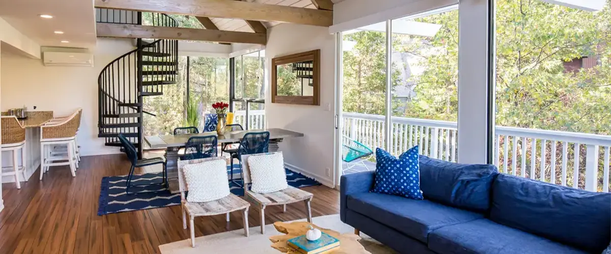 Modern open-plan living room with vaulted ceiling, wooden beams, spiral staircase, and large windows overlooking trees.