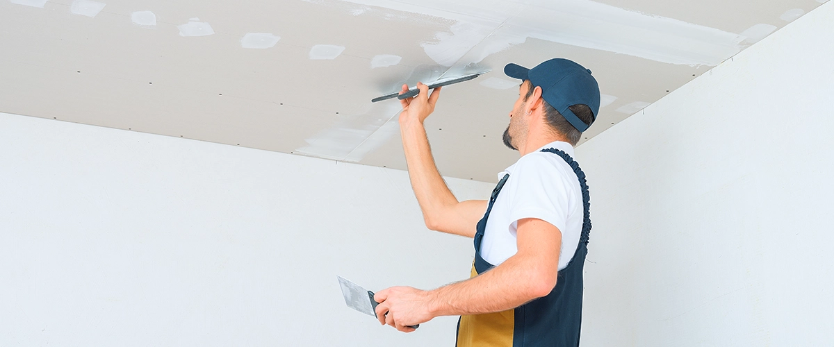 A uniformed worker applies putty to the drywall ceiling